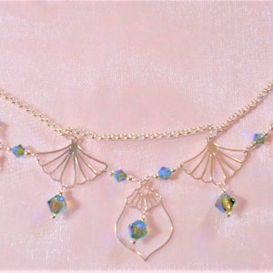 Può includere: Una collana d'argento con una catena delicata e più ciondoli. I ciondoli hanno la forma di ventagli e un design geometrico. La collana è adornata con cristalli blu iridescenti.