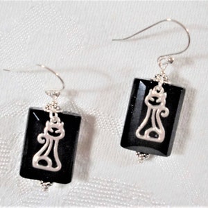 May include: A pair of black rectangular earrings with a silver cat design. The earrings have a faceted surface and are suspended from silver hooks. The cat design is a stylized silhouette.