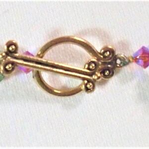 May include: A delicate gold clasp with a decorative design connects two strands of beaded jewelry. The beads are a mix of pink, green, and clear glass.
