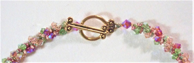 May include: A delicate gold clasp with a decorative design connects two strands of beaded jewelry. The beads are a mix of pink, green, and clear glass.