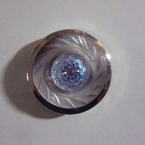 May include: A round, silver-toned button with a central blue, floral-patterned design. The button has a clear, textured center and a decorative, swirling pattern around the edge. The button is set against a white background.
