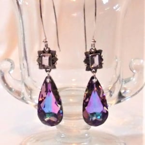 May include: A pair of dangle earrings with iridescent purple teardrop-shaped crystals. The earrings have a square, dark metal top with a smaller square crystal. The earrings are displayed in a clear glass vase.