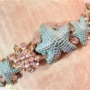 May include: A bracelet with light blue starfish and pink rhinestones. The starfish are detailed with small silver dots and are connected by gold-toned metal links. The bracelet is designed to be worn as a fashion accessory.