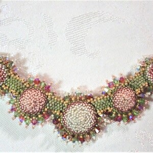May include: A beaded necklace with a floral design. The necklace features pink, green, and red beads arranged in a circular pattern. The necklace is made with a variety of beads, including seed beads, crystals, and pearls.