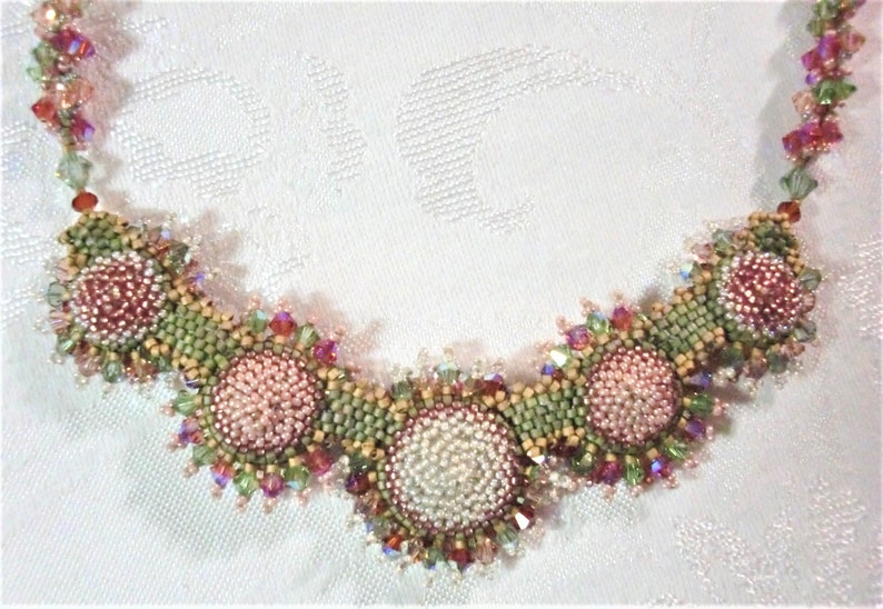 May include: A beaded necklace with a floral design. The necklace features pink, green, and red beads arranged in a circular pattern. The necklace is made with a variety of beads, including seed beads, crystals, and pearls.