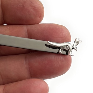 May include: A silver tie clip featuring a dinosaur design. The clip is attached to a black metal bar.