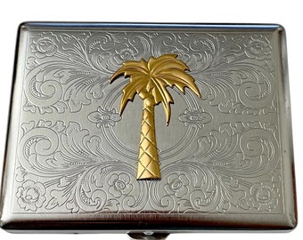 May include: Silver cigarette case with an engraved floral design and a gold palm tree on the front.