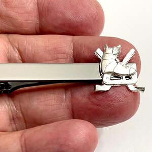 May include: Silver-toned tie bar featuring a hockey skate design. The tie clip has a rectangular bar with a shiny silver finish and a detailed hockey skate charm. A stylish accessory for hockey enthusiasts.