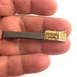 Pode incluir: Um prendedor de gravata dourado com uma caixa de correio em miniatura com a inscrição "US MAIL". O prendedor é preso a uma barra de metal marrom escuro.