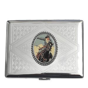 May include: A silver metal cigarette case with an embossed diamond pattern. The center features an oval image of a cowboy on a horse, framed by a rope design. The case has a hinged lid and a rectangular shape.