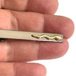 Può includere: Un fermacravatta argentato con un design a serpente dorato. Il fermacravatta rettangolare presenta un motivo a serpente strutturato e dettagliato su un'estremità. Il design del serpente è a forma di onda.