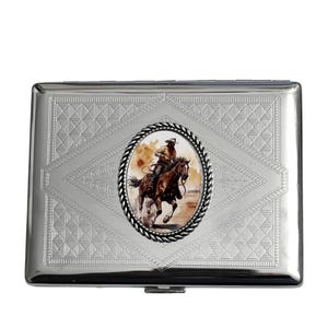 May include: A silver cigarette case with a detailed embossed pattern. The center features an oval image of a cowboy riding a horse. The image is surrounded by a black rope-like border. The case has a rectangular shape with a hinged opening.