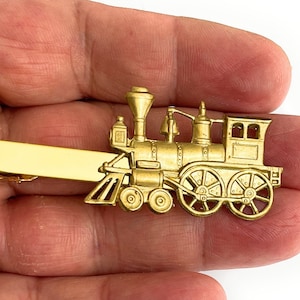 Puede incluir: Un pasador de corbata dorado con forma de tren de vapor antiguo. El pasador presenta ruedas detalladas, una campana y una chimenea. El tren está unido a una barra rectangular para sujetarlo a una corbata.