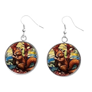 May include: A pair of round earrings featuring a brown squirrel in a forest scene. The squirrel is set against a backdrop of trees, water, and colorful sky. The earrings have silver-colored hooks.
