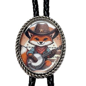 May include: Oval bolo tie with a cartoon fox wearing a cowboy hat and playing a banjo. The fox has orange fur and is wearing a blue shirt and red bandana. The tie has a silver-colored rope border and black braided cord.
