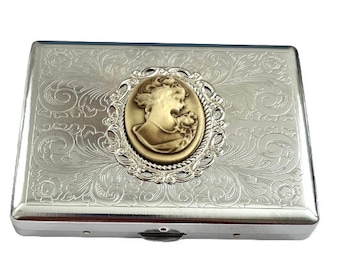 May include: Silver cigarette case with an ornate floral design and a cameo of a woman's profile in a gold frame.
