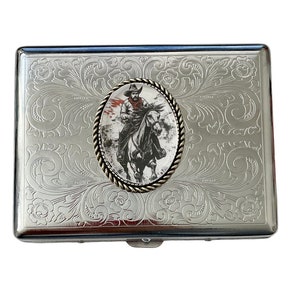 May include: Silver cigarette case with an engraved floral design and a round, silver-toned medallion featuring a black and white image of a cowboy riding a horse.