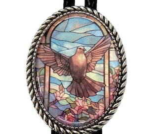 May include: Oval-shaped bolo tie with a silver-toned rope-like border. The centerpiece features a stained-glass design of a dove with outstretched wings, set against a backdrop of blue sky and pink flowers. The tie has a black braided cord.