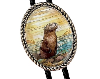 May include: A bolo tie with a silver oval pendant featuring a painted image of an otter looking up at the sky. The pendant has a rope-like border. The bolo tie has black cords.