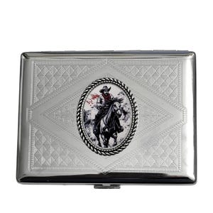 May include: A silver metal cigarette case with a decorative diamond pattern. The case features an oval image of a cowboy on a horse. The cowboy is wearing a hat and red scarf. The image is surrounded by a braided border.