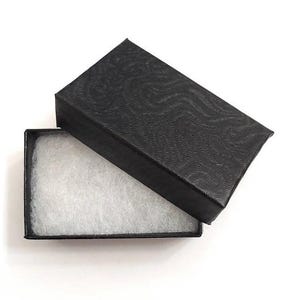 Op de afbeelding: Een open zwarte geschenkdoos met een getextureerd deksel en een witte, donzige binnenkant. De rechthoekige doos staat tegen een witte achtergrond. Het deksel heeft een subtiel golfpatroon.