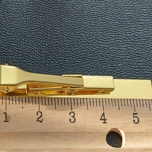 May include: Gold tie clip measuring 5 cm in length. The clip is a simple design with a straight bar and a spring-loaded clasp.