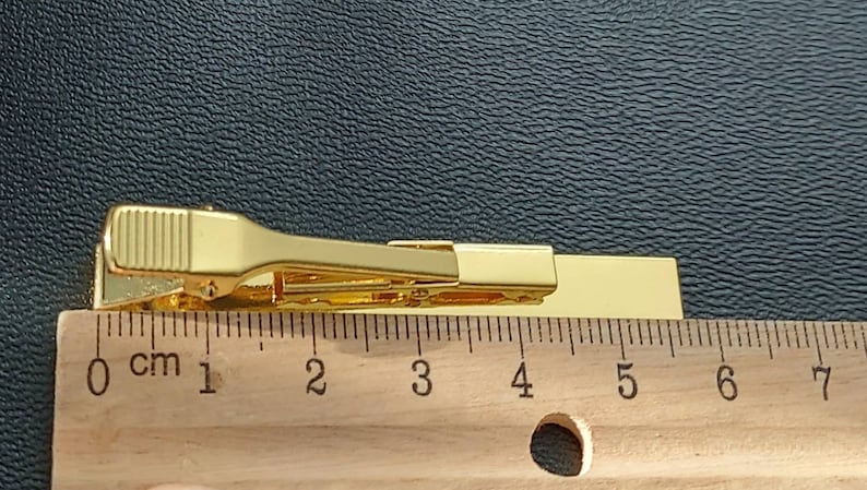 May include: Gold tie clip measuring 5 cm in length. The clip is a simple design with a straight bar and a spring-loaded clasp.