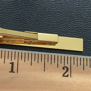 May include: A gold-colored tie bar measuring approximately 2 inches in length. The tie bar is shown next to a ruler for size reference.