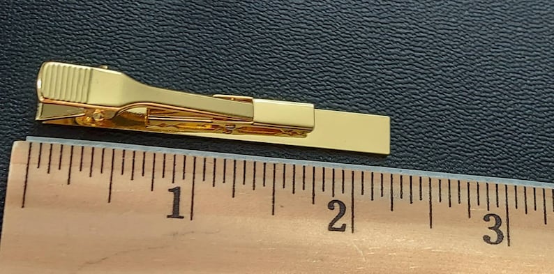 May include: A gold-colored tie bar measuring approximately 2 inches in length. The tie bar is shown next to a ruler for size reference.
