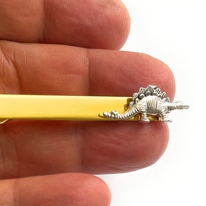 May include: A gold-toned tie slide featuring a small, silver-coloured stegosaurus dinosaur figure attached to the right side. The tie slide has a smooth, polished finish and a rectangular shape, providing a secure clip mechanism for fastening a necktie.