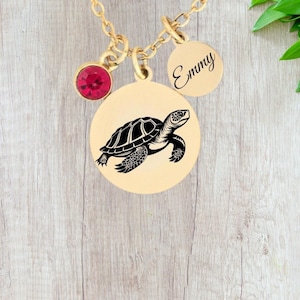 Puede incluir: Collar dorado con un colgante con un diseño de tortuga marina negra. También se adjuntan una gema roja y un dije redondo con el nombre "Emmy". El collar se exhibe sobre una superficie de vetas de madera clara.