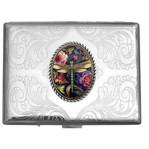 May include: A rectangular silver-toned metal case featuring an intricate engraved scrollwork pattern across the surface. Centered on the lid is an oval inlay framed by a twisted rope border, displaying a colorful dragonfly over floral imagery.