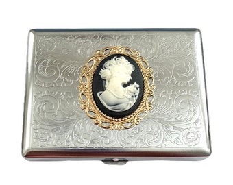 May include: A silver cigarette case with an ornate floral design and a cameo of a woman in a gold frame.