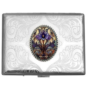 May include: A rectangular silver-toned metal case featuring an intricate engraved scrollwork pattern across its surface. Centered on the lid is an oval inlay framed by a twisted rope border, displaying a colorful illustration of a purple flower.