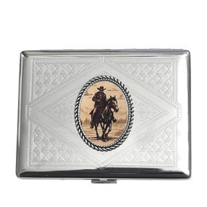 May include: A silver metal cigarette case with a decorative diamond pattern. An oval image of a cowboy on a horse is centered on the case. The case has a hinged lid and a clasp closure.