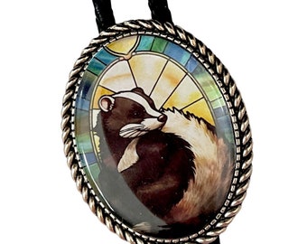 May include: A black bolo tie with a silver oval pendant featuring a stained glass image of a badger. The pendant has a rope-like border.