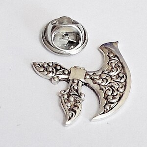 May include: A silver-colored metal axe pin with intricate floral detailing. The pin has a separate backing piece for attachment. The axe head features a curved blade and a detailed handle, perfect for adding a unique touch to clothing or accessories.