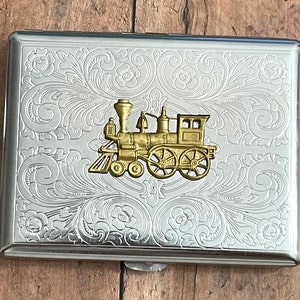 May include: A silver cigarette case with an ornate floral design and a gold-colored train engine on the front.