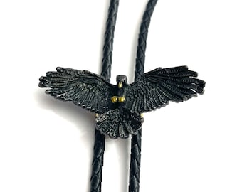 May include: Black bolo tie featuring a detailed metal eagle pendant. The eagle has its wings spread wide and is facing forward. The bolo tie is made of black leather cord.