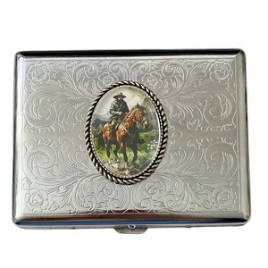 May include: Silver cigarette case with an engraved floral design and a round, metal inlay featuring a picture of a cowboy riding a horse.