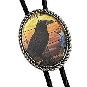 May include: A black bolo tie. The silver-toned oval pendant features a raven and a blue bird against a stained-glass background, framed by a twisted rope design. The tie is made of black braided leather.