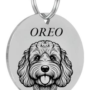 May include: A round, silver pet tag with a detailed black illustration of a dog's face. The name "OREO" is printed in black above the dog's face. A silver ring is attached for easy attachment to a collar.