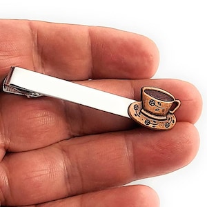 Może przedstawiać: Srebrny klips do krawata z dekoracyjnym, brązowym kubkiem do herbaty i spodkiem. Filiżanka jest wypełniona ciemnobrązowym płynem i ma kwiatowe detale. Klips jest trzymany w dłoni.