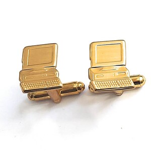 May include: A pair of gold-colored cufflinks shaped like vintage computer monitors. Each cufflink features a screen, keyboard, and base, with a connecting bar and a rounded clasp. The cufflinks are set against a white background.