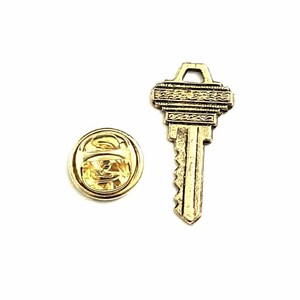 Puede incluir: Un pin con forma de llave dorada y un respaldo redondo se muestran sobre un fondo blanco. El pin de la llave tiene detalles intrincados y una superficie texturizada. El respaldo del pin tiene un diseño simple y redondo.
