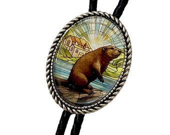 May include: A black bolo tie with a silver oval pendant featuring a cartoon beaver standing on a rock by a river. The background is a stained glass style image of a house and a sunrise.
