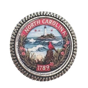 Op de afbeelding: Een ronde, zilverkleurige pin met het zegel van de staat North Carolina. Het zegel toont een vuurtoren, oceaangolven, een rode vogel en de tekst "NORTH CAROLINA" en "1789". De pin heeft een getextureerde rand.