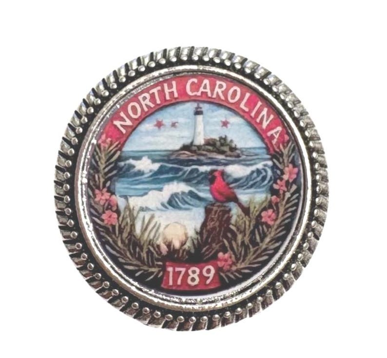 Op de afbeelding: Een ronde, zilverkleurige pin met het zegel van de staat North Carolina. Het zegel toont een vuurtoren, oceaangolven, een rode vogel en de tekst "NORTH CAROLINA" en "1789". De pin heeft een getextureerde rand.
