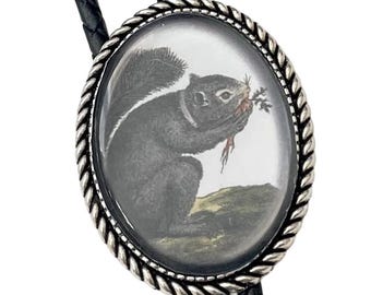 May include: A black bolo tie with a silver oval pendant featuring a black and white illustration of a squirrel eating a carrot.