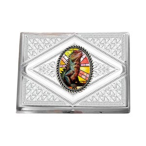 Op de afbeelding: Een zilveren sigarettenetui met een decoratief geometrisch patroon. Het midden toont een ovaal beeld van een hagedis in glas-in-loodstijl, met rode, oranje, gele en groene tinten. De etui heeft een rechthoekige vorm.